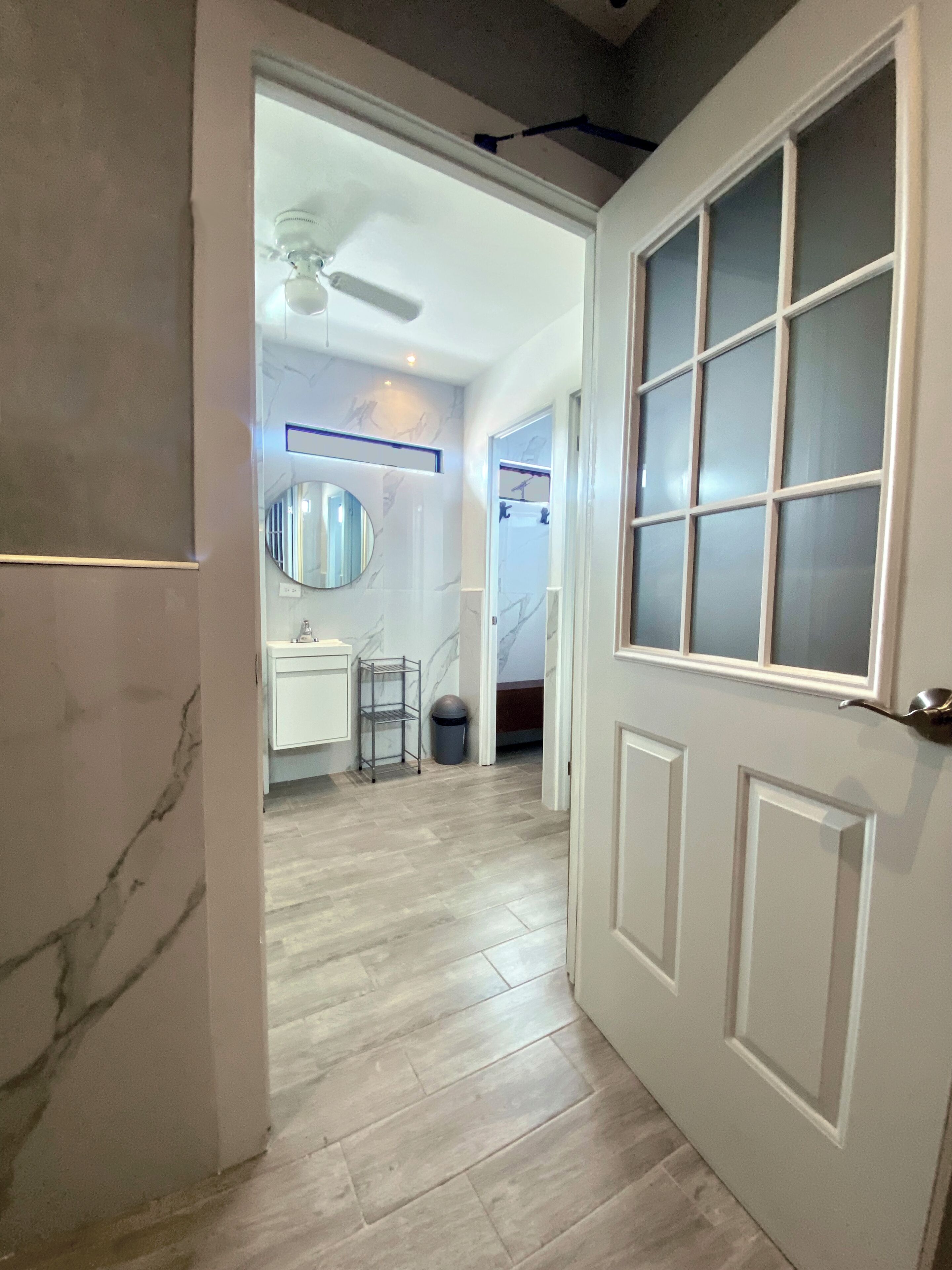 A hallway with a white door, a ceiling fan, and a view into a room with a bathroom.