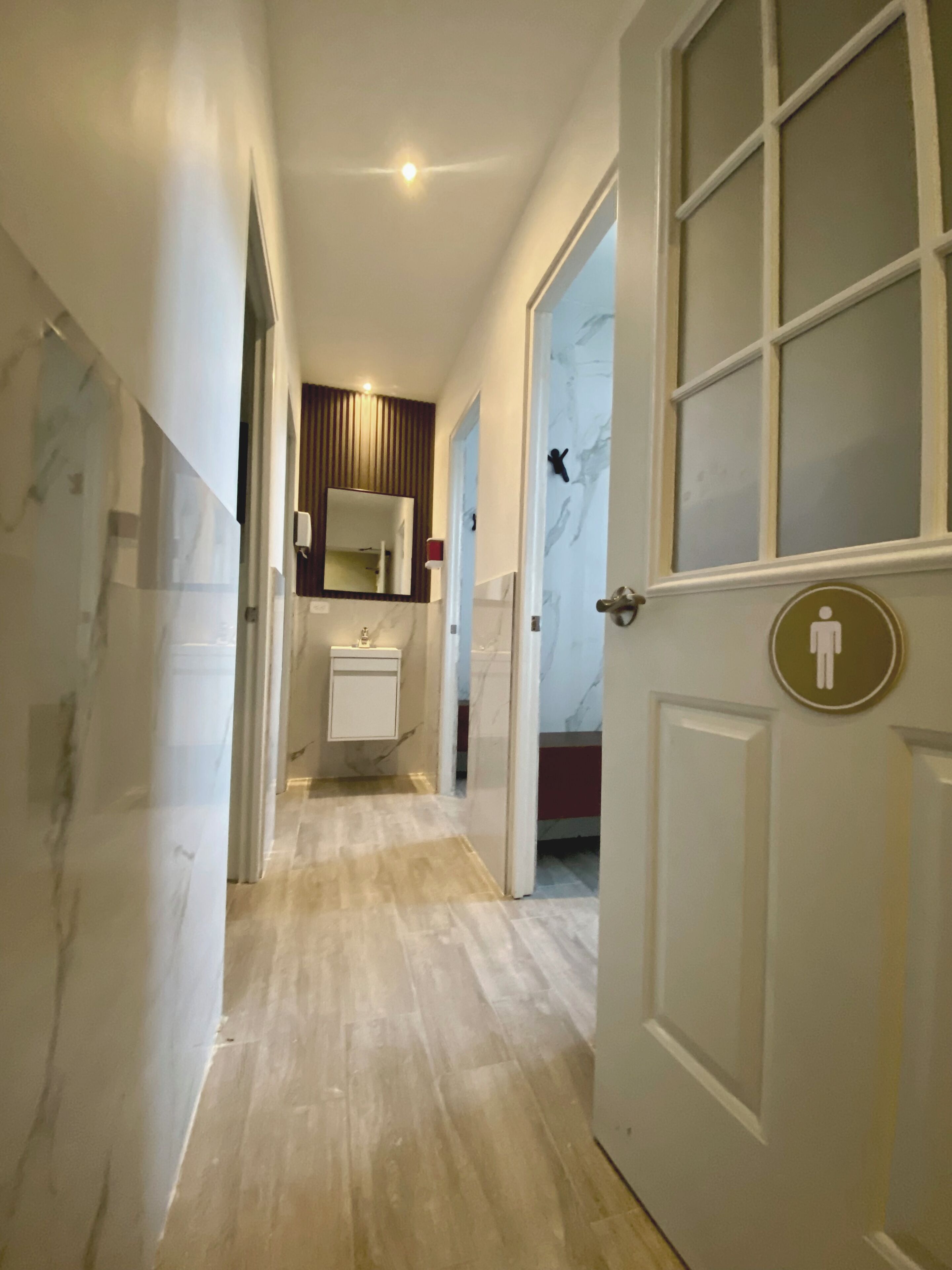 A hallway with a marble floor, a white door with a restroom sign, and a mirror above a sink.