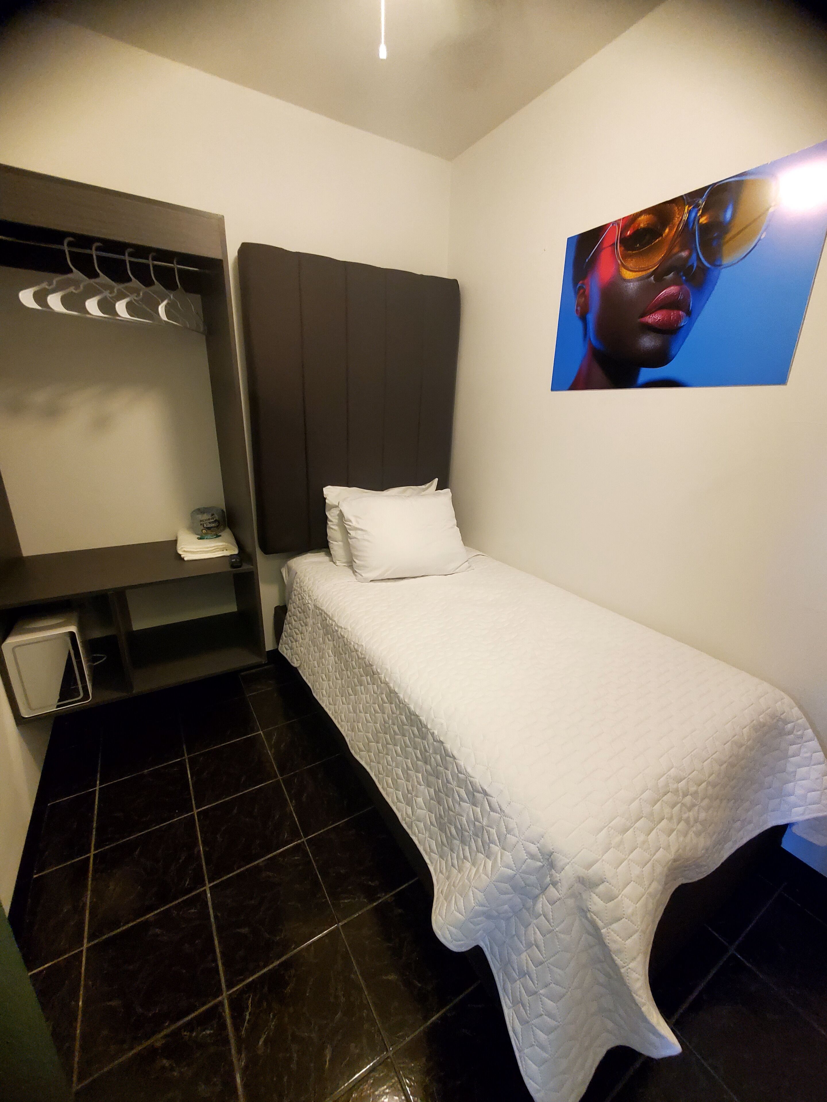 Una habitación de hotel pequeña y ordenada con una cama, una mesita de noche, un espejo y un cuadro colgado en la pared.
