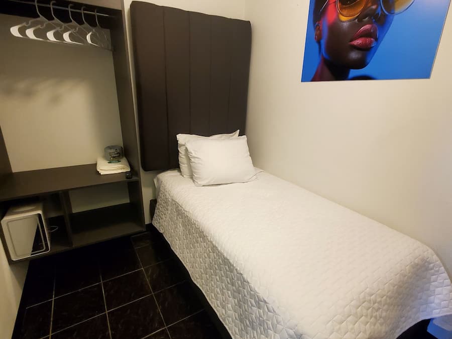 Una habitación de hotel pequeña y ordenada con una cama, una mesita de noche, un espejo y un cuadro colgado en la pared.