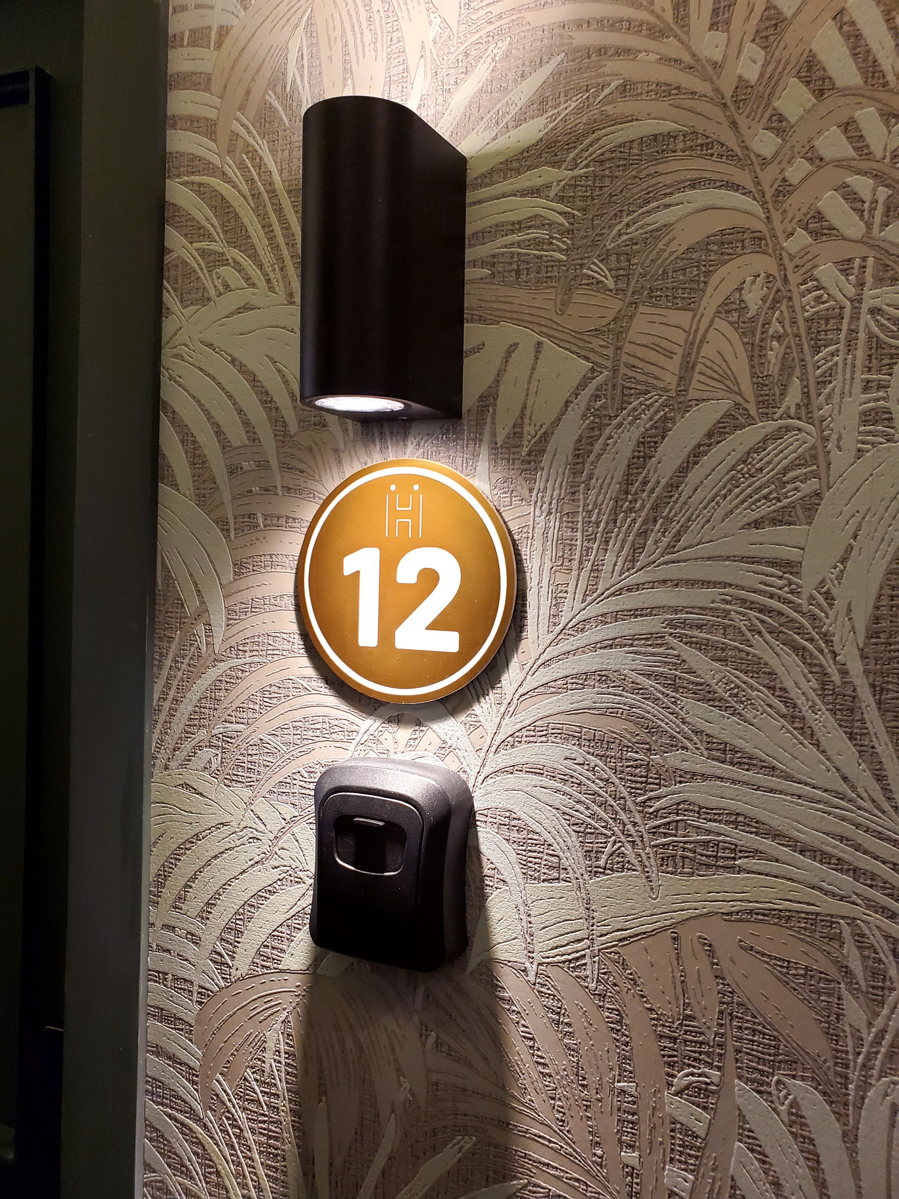 A wall-mounted light fixture, a circular sign with the number 12, and a small black object on a textured wall.
