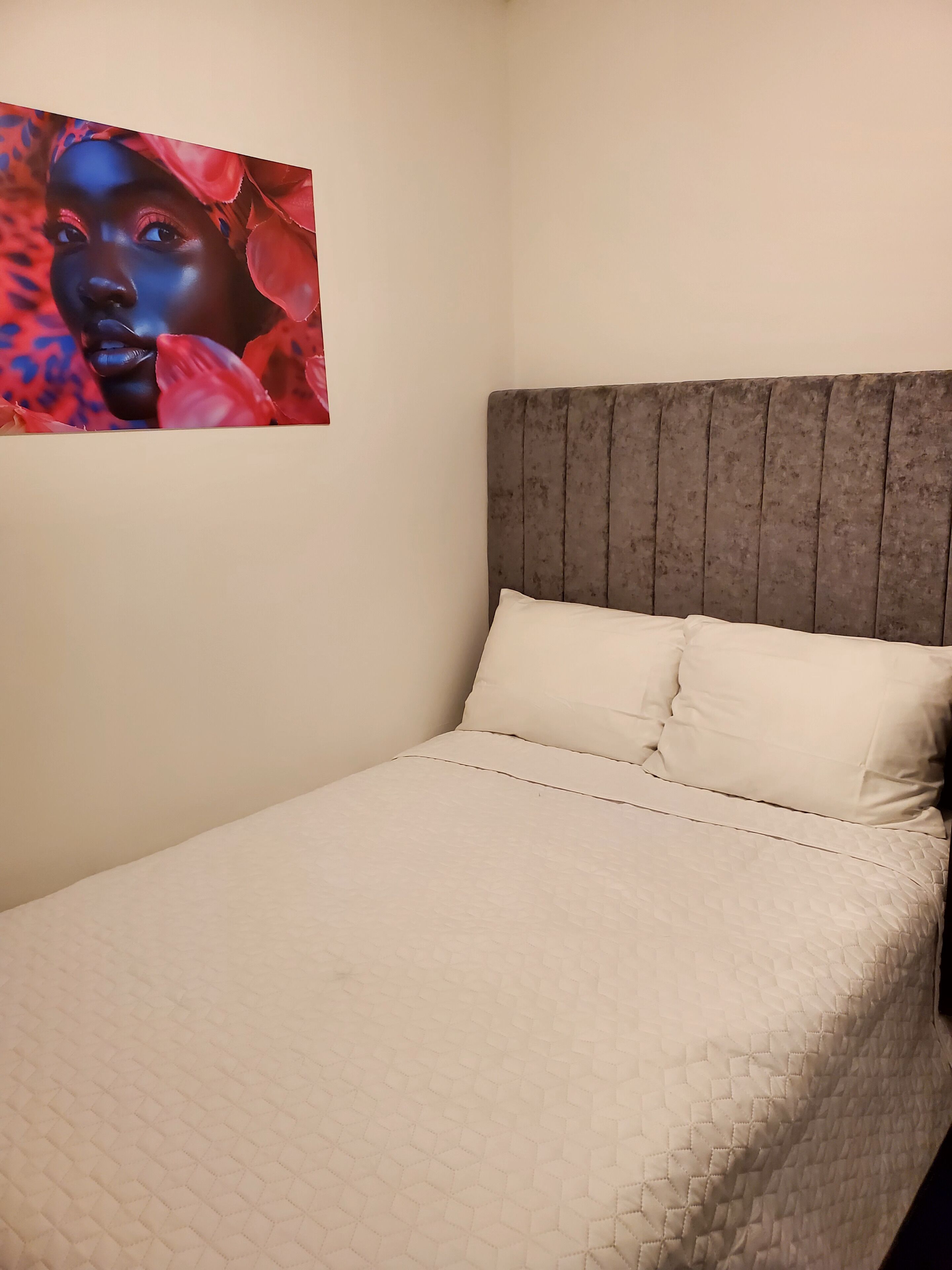 A bed with a quilted cover, a grey upholstered headboard, and a wall with a framed picture.
