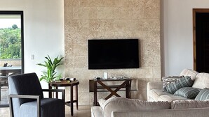 A spacious living room with a stone wall, a ceiling fan, a flat-screen TV, a sofa, a chair, and a small table with a plant. - Panoramic Lake Arenal Sunset View Villa with AC (Nuevo Arenal)