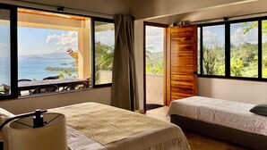 A hotel room with a bed, a bedside table, and a view of a coastal landscape. - Panoramic Lake Arenal Sunset View Villa with AC (Nuevo Arenal)