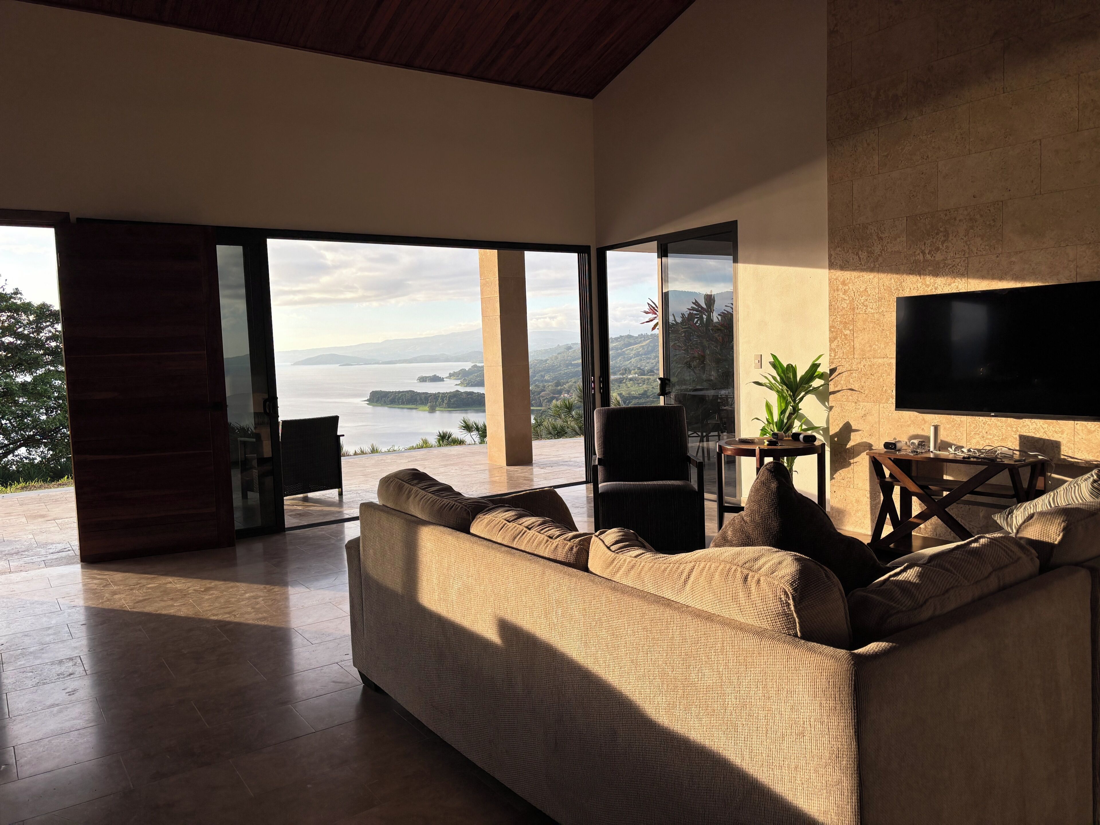 Ein modernes Wohnzimmer mit einer großen Couch, einem Couchtisch und einem Flachbildfernseher. Das Zimmer verfügt über große Glasfenster mit Blick auf ein Gewässer und Hügel.