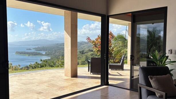A modern interior with a large glass door leading to a balcony with a view of a coastal landscape. - Panoramic Lake Arenal Sunset View Villa with AC (Nuevo Arenal)
