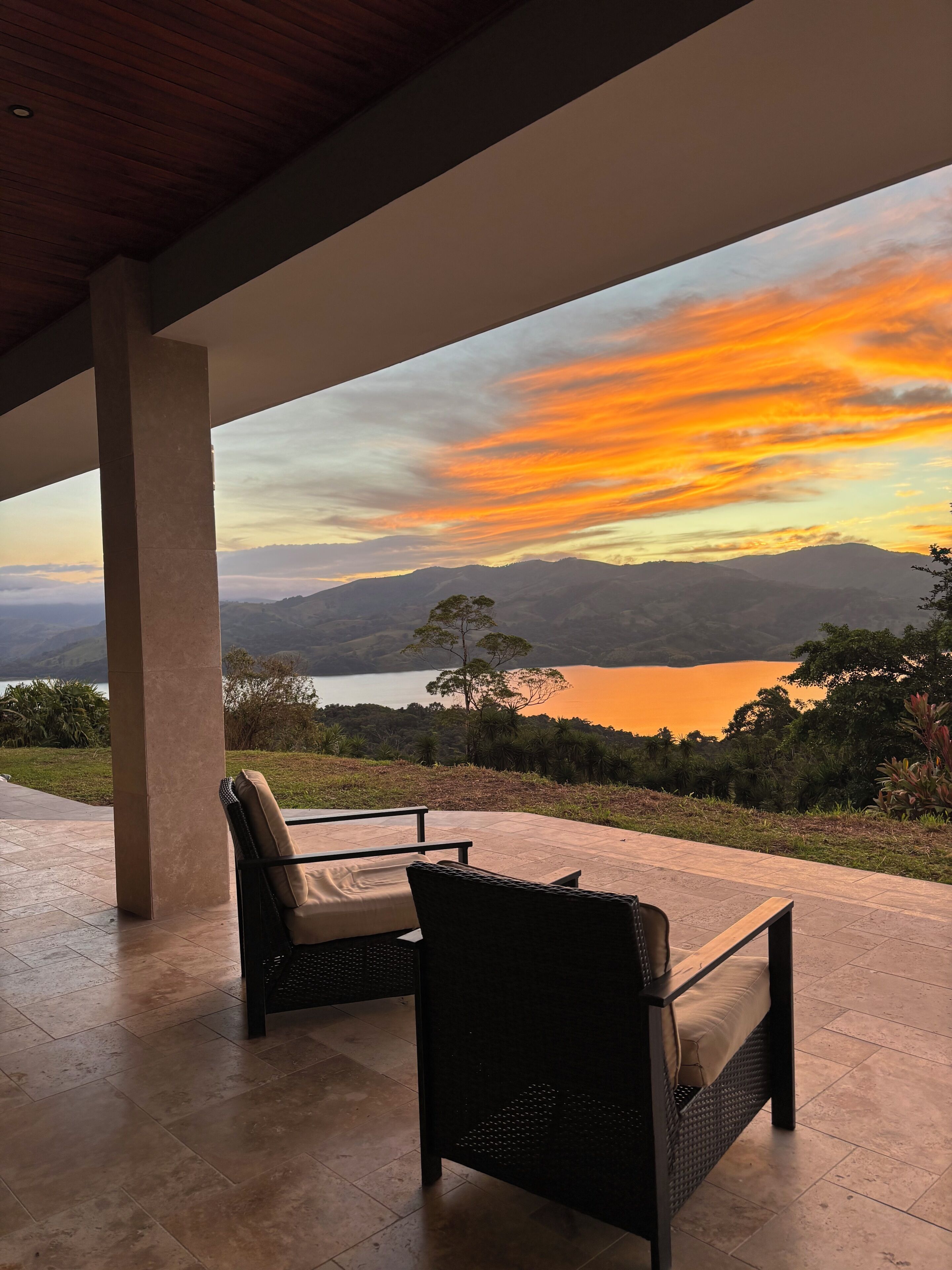 Un espacio de estar al aire libre cubierto con dos sillones de mimbre y una mesa, con vistas a un lago y montañas durante el atardecer.