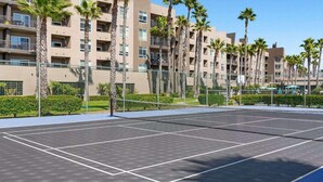 A tennis court with a net, surrounded by a fence and palm trees, with a multi-story building in the background.
