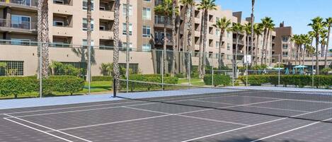 A tennis court with a net, surrounded by a fence and palm trees, with a multi-story building in the background.