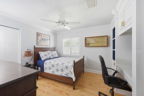 Une chambre à coucher comprenant un lit, un bureau, une chaise, un ventilateur de plafond et une fenêtre avec des stores.