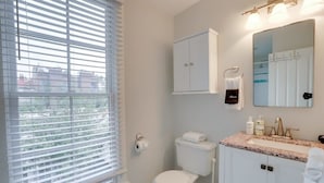 Et badeværelse med toilet, vask, spejl, skab og et vindue med persienner.