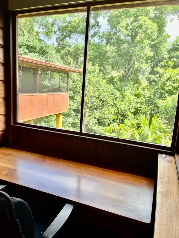 A room with a large window, wooden desk, and a view of a wooden deck and lush greenery.