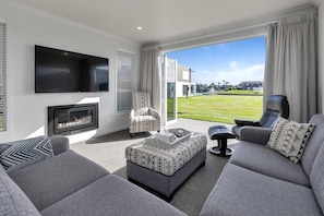 A modern living room with a flat-screen TV, fireplace, comfortable seating, and a large window with a view of a golf course. - Waterfront Base with Private Pontoon and Kayaks (One Tree Point)