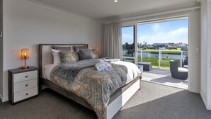 A bedroom with a large bed, a nightstand, a lamp, and a view of a balcony with outdoor furniture. - Waterfront Base with Private Pontoon and Kayaks (One Tree Point)