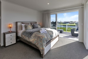 A bedroom with a large bed, a nightstand, a lamp, and a view of a balcony with outdoor furniture. - Waterfront Base with Private Pontoon and Kayaks (One Tree Point)