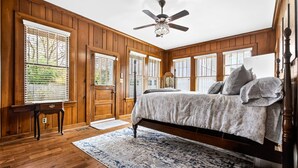 A bedroom with a wooden floor, a bed with pillows, a ceiling fan, and large windows with blinds. - Historic Midtown Brick Home with Fire Pit (Columbus)