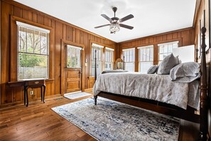 Ein Schlafzimmer mit Holzboden, einem Bett mit Kissen, einem Deckenventilator und großen Fenstern mit Jalousien.
