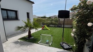 Un jardin à l’arrière d’une maison, avec une terrasse, un petit espace de jeux et un panier de basketball.