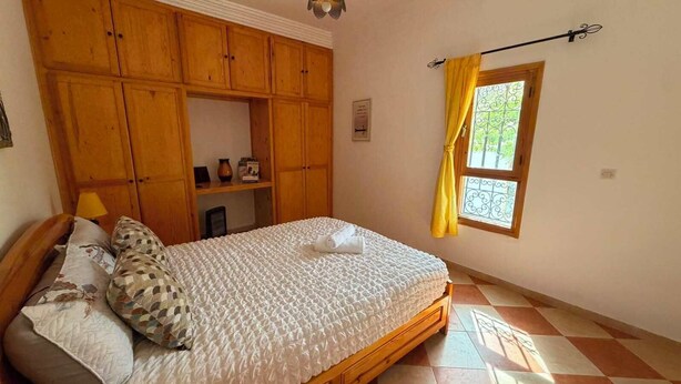 A bedroom with a bed, wooden wardrobe, a window with yellow curtains, and a tiled floor.