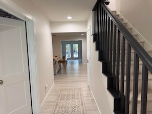 A hallway with a wooden floor, a patterned rug, a staircase with black railings, and a door on the left. - Weldons' Kyle Field Cottage (College Station)