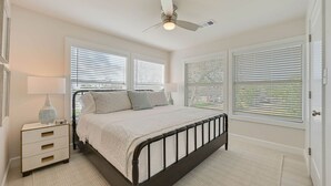 Un dormitorio con cama, mesitas de noche, lámpara, ventilador de techo y ventanas amplias con persianas.