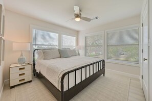 A bedroom with a bed, bedside tables, a lamp, a ceiling fan, and large windows with blinds. - Weldons' Kyle Field Cottage (College Station)