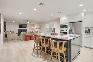 A modern kitchen with a marble island, wooden bar stools, and stainless steel appliances. - Weldons' Kyle Field Cottage (College Station)