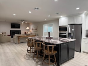 A modern kitchen with a central island, bar stools, a refrigerator, and a dining area with a sofa and table. - Weldons' Kyle Field Cottage (College Station)