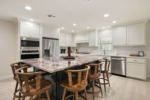 A modern kitchen with white cabinetry, stainless steel appliances, a marble island, and wooden bar stools. - Weldons' Kyle Field Cottage (College Station)