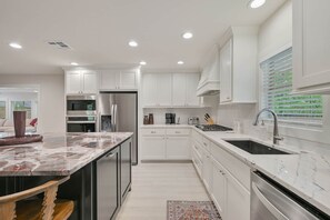 A modern kitchen with white cabinetry, stainless steel appliances, and a marble countertop. - Weldons' Kyle Field Cottage (College Station)