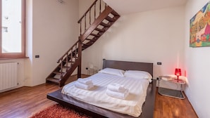 A bedroom with a wooden staircase, a bed with white linens, a red lamp on a side table, and a colorful painting on the wall. - Venezia Terrace (Florence)