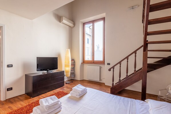 A bedroom with a bed, a TV on a dresser, a window, a radiator, and a wooden staircase. - Venezia Terrace (Florence)