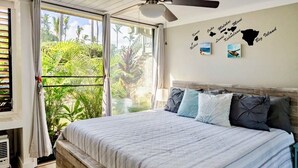 A bedroom with a large bed, a ceiling fan, and a view of tropical vegetation through a sliding door. - Cozy 1-bedroom condo at Turtle Bay, North Shore AC, private lanai, full kitchen. (Kahuku)
