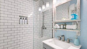 A bathroom with white subway tiles, a glass-enclosed shower, a vanity with a white sink, and a mirror with shelves above. - Lido Bungalow Walk to Beach & St. Armand's Circle (Sarasota)
