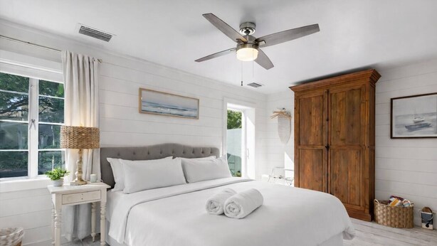 Um quarto com cama, ventoinha no teto, janela com cortinas, guarda-roupa de madeira e um quadro na parede.