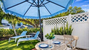 Um pátio com uma mesa posta para dois, um guarda-sol azul e uma cerca branca com plantas.