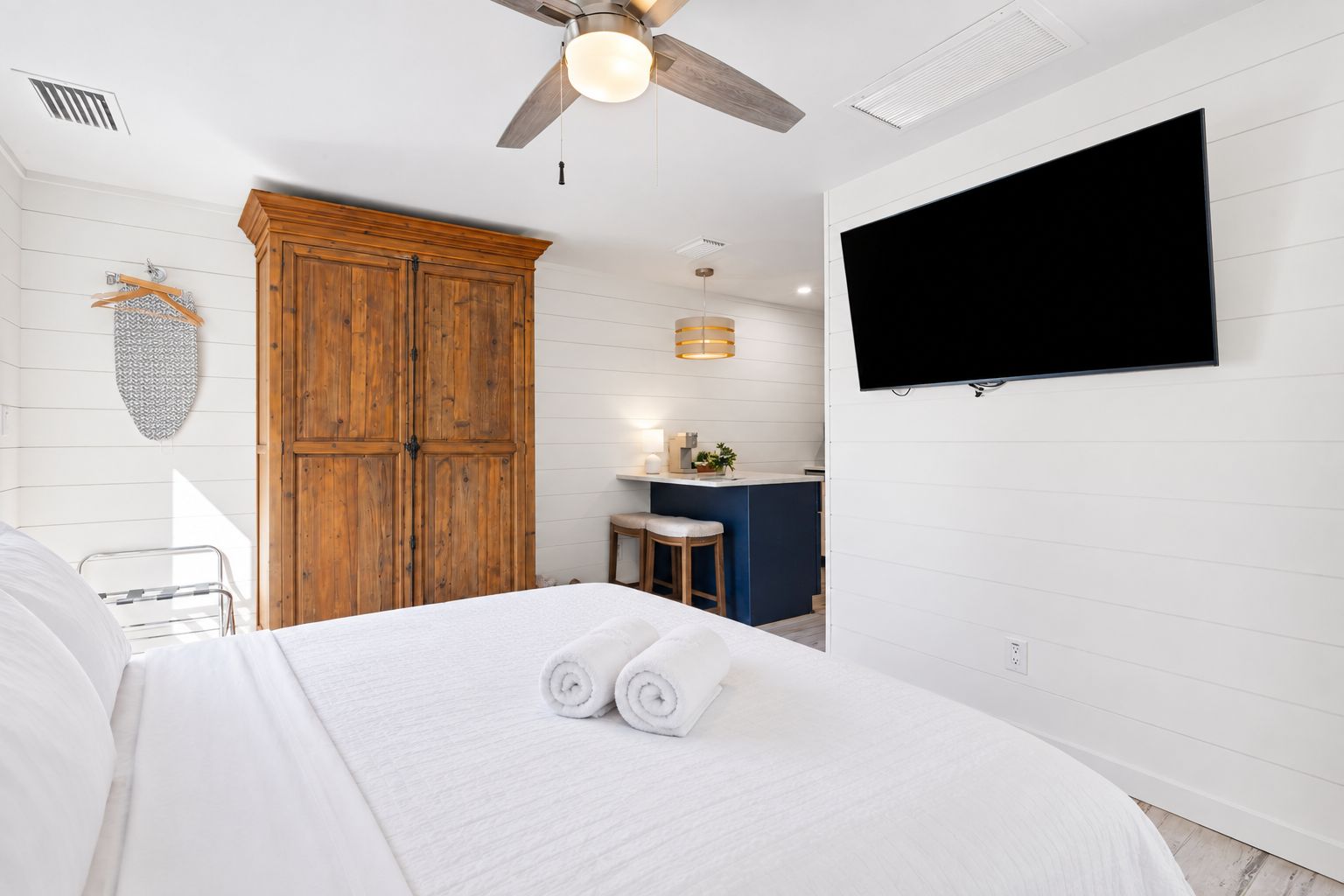 A hotel room with a bed, a wooden wardrobe, a TV mounted on the wall, and a small kitchenette area.