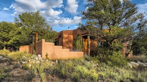 A rustic adobe-style building with a wooden pergola, surrounded by native vegetation and rocks.