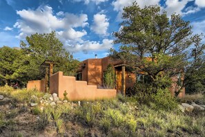 A rustic adobe-style building with a wooden pergola, surrounded by native vegetation and rocks.