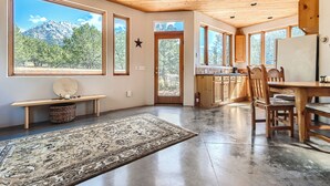 A spacious kitchen with wooden cabinets, a dining table, and chairs. Large windows offer views of a mountain and trees.