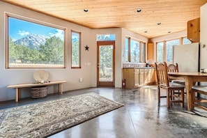 A spacious kitchen with wooden cabinets, a dining table, and chairs. Large windows offer views of a mountain and trees.
