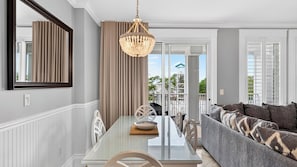 A dining area with a glass table, chairs, a chandelier, and a sofa with patterned cushions. - Wharf & Wander by Avantstay Walk to Village + Beach Tram + Resort Pool + Golf Baytowne Wharf (Miramar Beach)