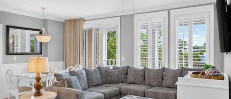 A living room with a sectional sofa, a dining table with chairs, a ceiling fan, and large windows with shutters.