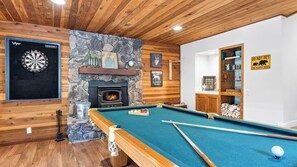 A billiards table with a pool cue, a dartboard, and a fireplace with a stone surround. - London Pines Lodge by Avantstay Pine-view Big Bear Cabin Hot Tub + Game Room (Big Bear City)