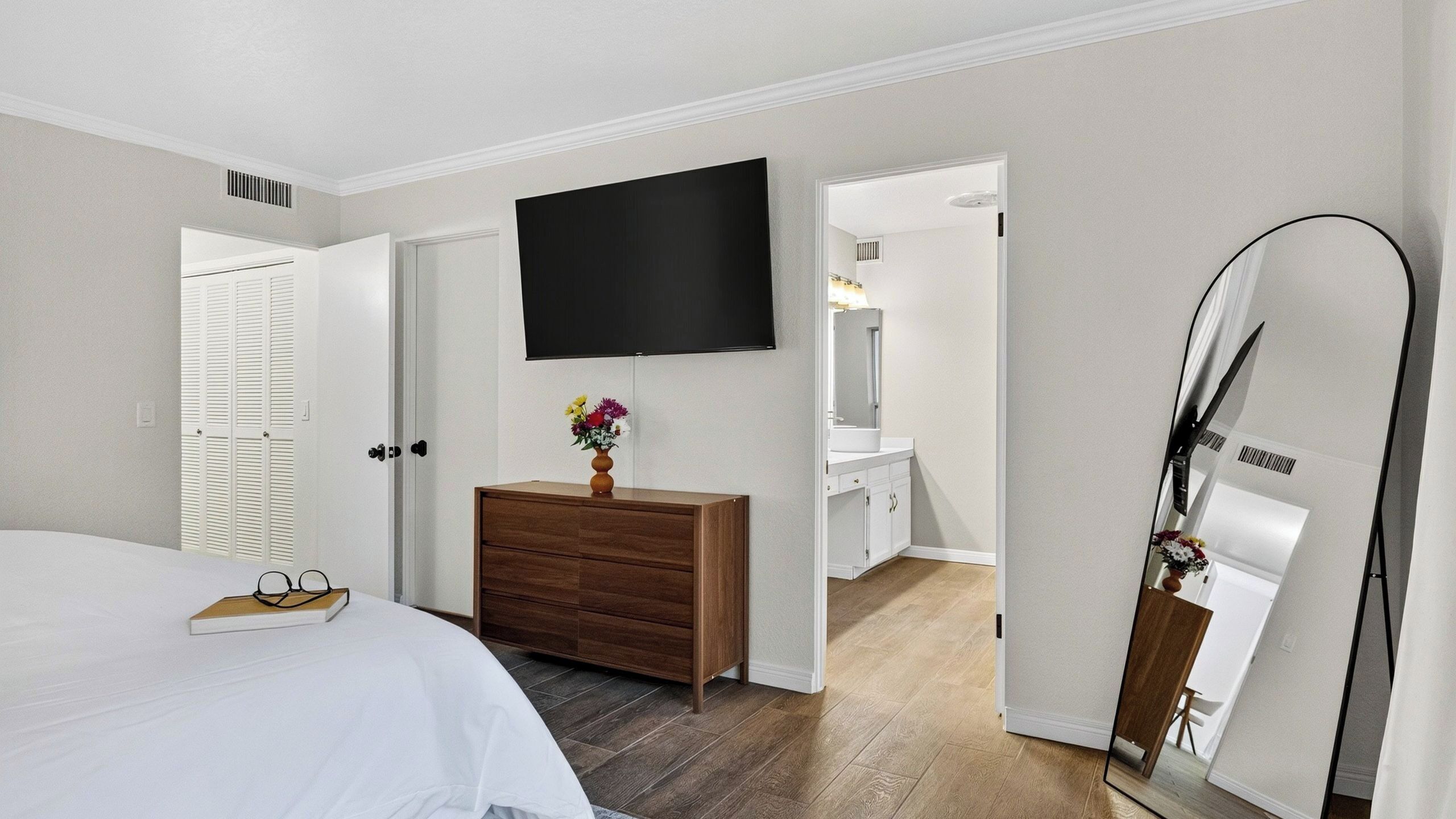 Un dormitorio con cama, tocador, televisor, espejo y baño visible a través de una puerta abierta.