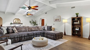 Ein modernes Wohnzimmer mit einem Ecksofa, einem runden Hocker, einem Deckenventilator und einem Bücherregal.