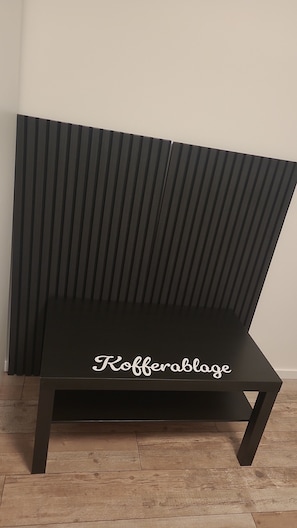 Un banc en métal noir avec un dossier à lattes et l’inscription « Kofferablage » sur l’assise.