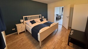 Quarto com cama, mesas de cabeceira, abajur, guarda-roupa e porta que dá para uma sala de jantar.