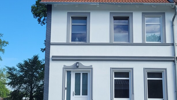 Uma casa de dois andares com painéis solares no telhado, uma porta branca e um pequeno jardim com degraus que levam à entrada.