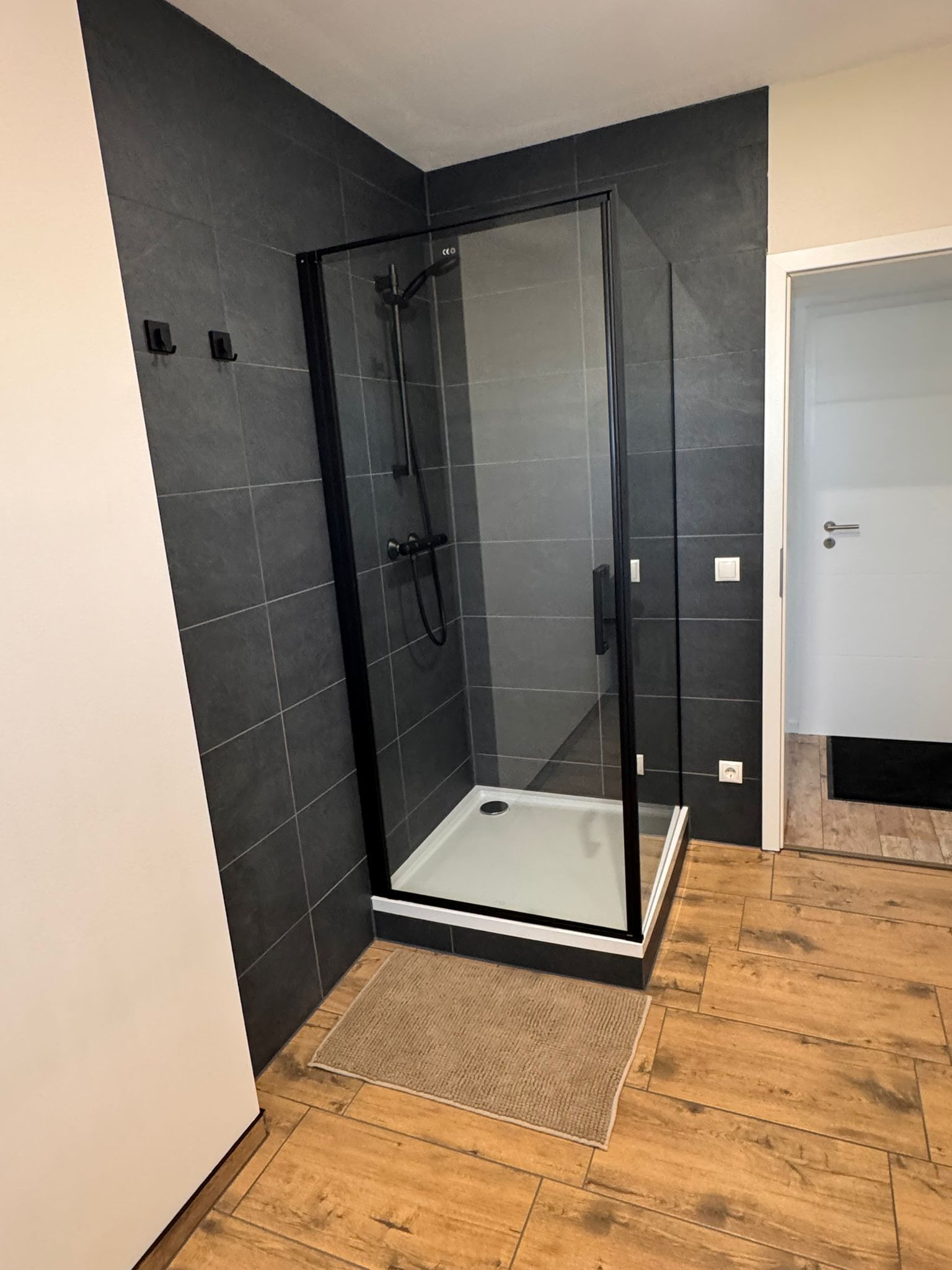 Ein modernes Badezimmer mit einer in Glas eingefassten Dusche, dunklen Fliesenwänden und einem Holzboden.